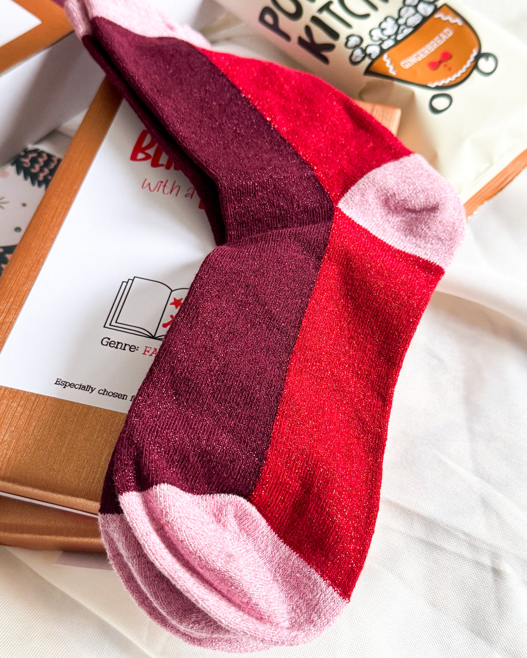 A close-up of a crew sock with color-blocked stripes in dark burgundy, bright red, and light pink, resting on a white fabric surface next to a cardboard box and a branded tote bag.