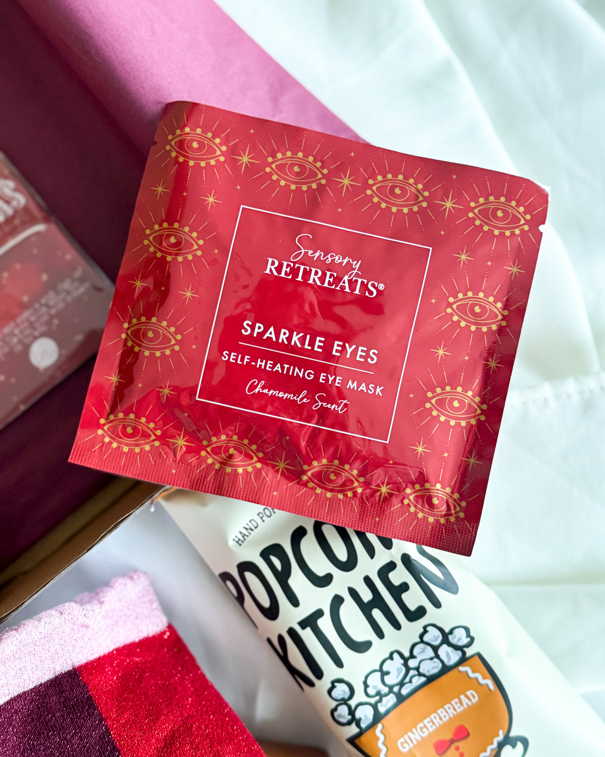A red Sensory Retreats Sparkle Eyes self-heating eye mask packet with gold foil eye illustrations, shown alongside a white Popcorn Kitchen snack bag and a pink plush item in a gift box setting.