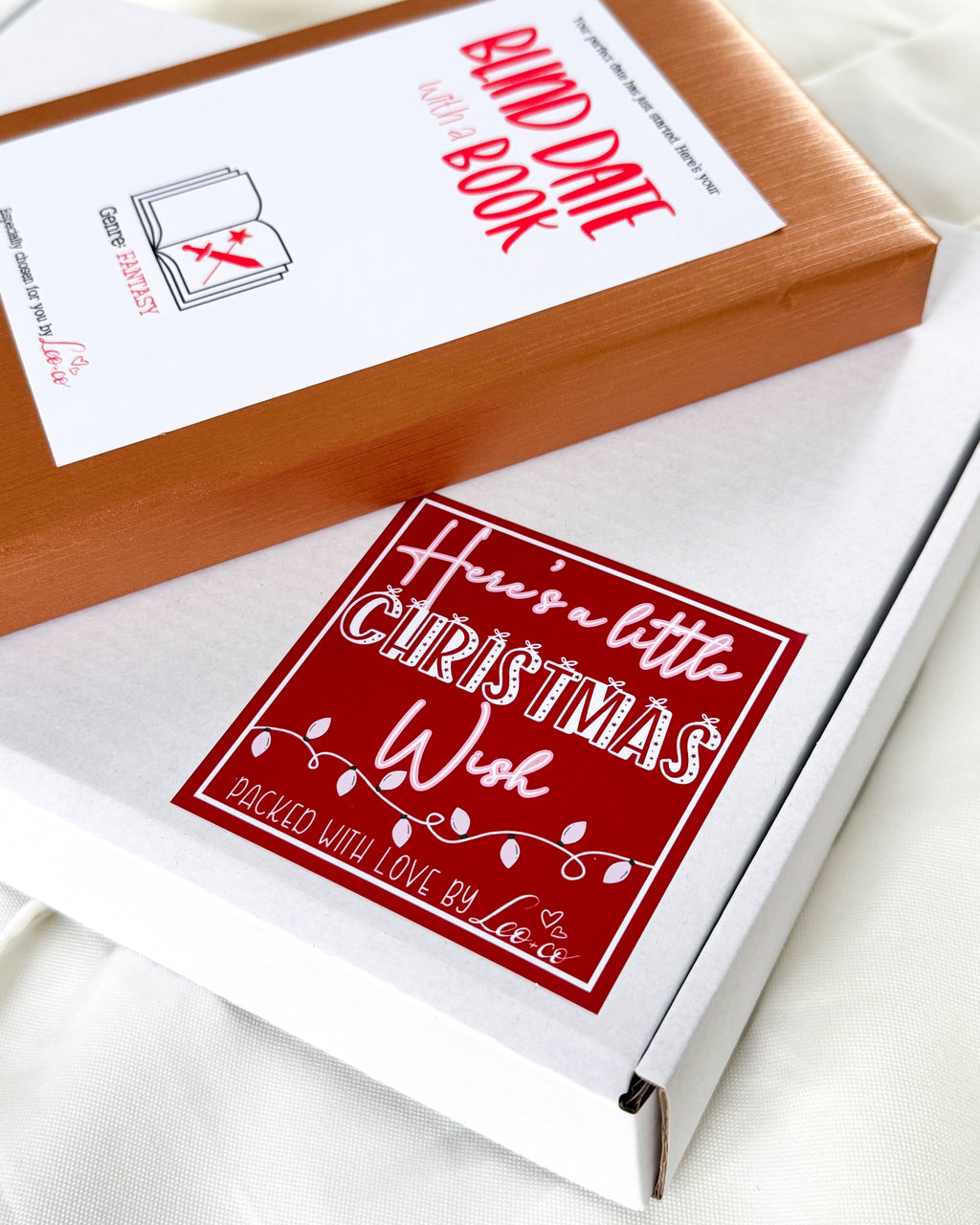 A white gift box with a red and white 'Here's a little Christmas Wish' sticker, alongside a brown box labeled 'Bind Date Book' by Leo + CO Gifts  on a white fabric background.