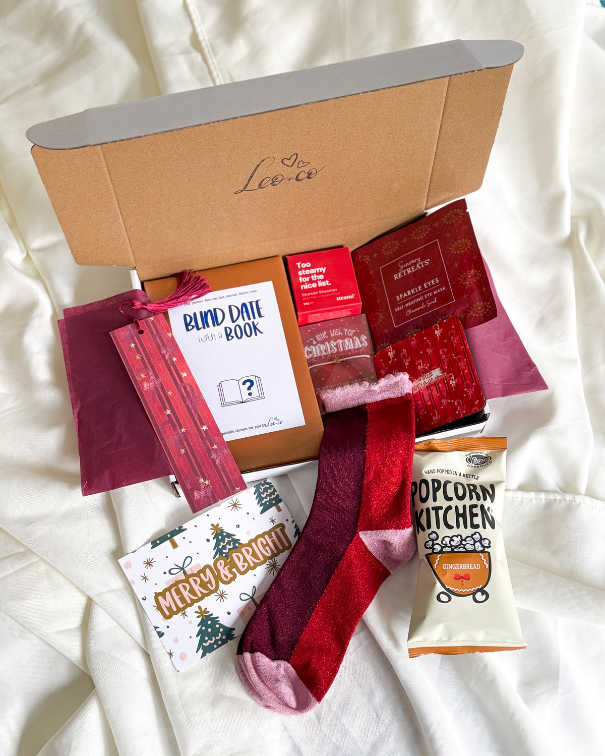 Christmas Blind Date With a Book gift box by Leo + Co, featuring a mystery book, cosy socks, shower steamer, Christmas wish bracelet, festive coaster, bookmark, self-heating eye mask and vegan gingerbread popcorn.
