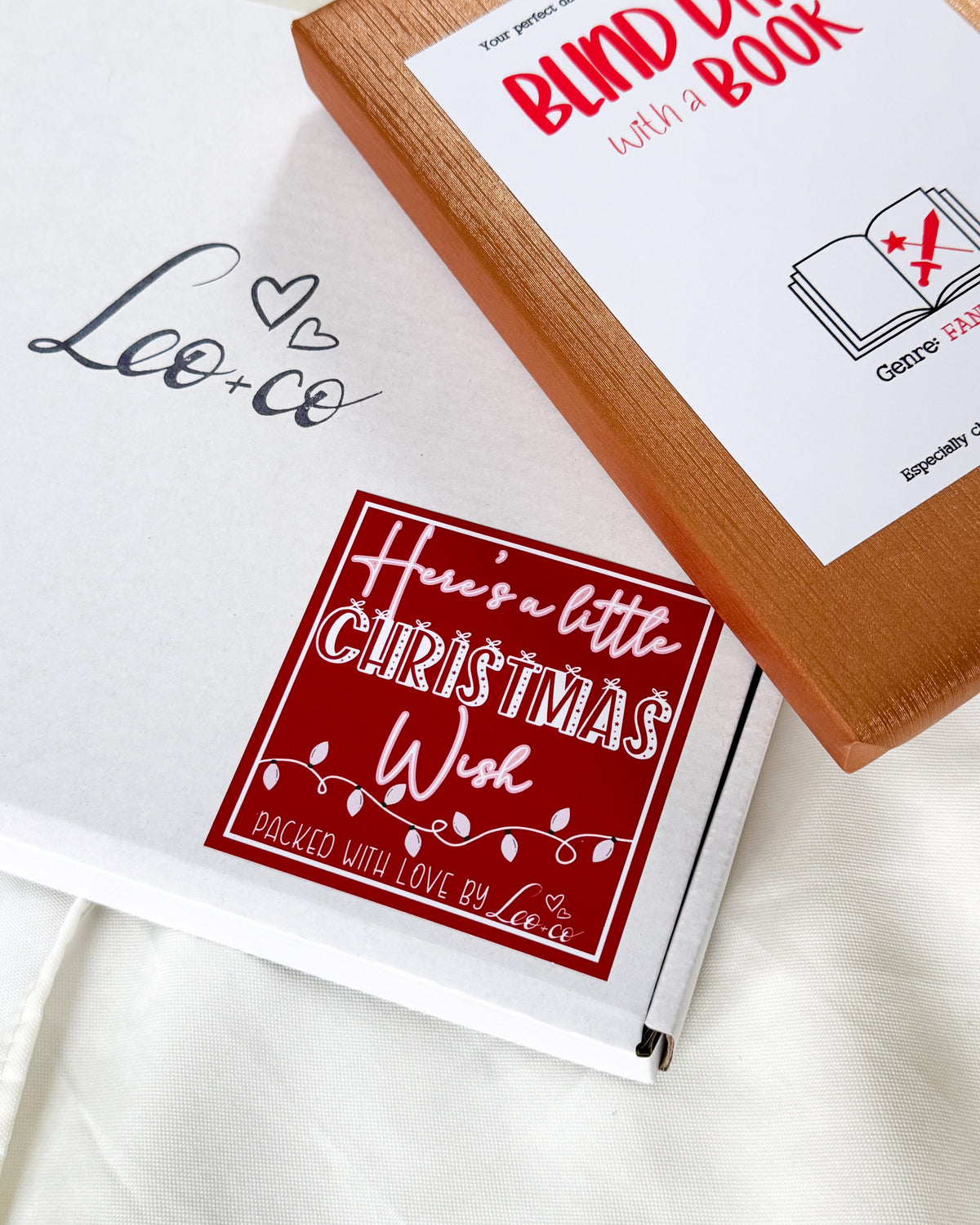 A white gift box with a red square sticker reading 'Here's a little Christmas Wish' and a wooden book package labeled 'Blind Date with a Book' placed on top, on a white fabric background.