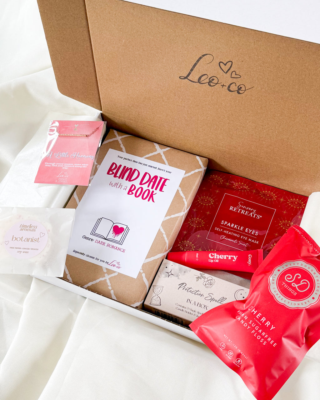 An open kraft cardboard gift box labeled 'Leo co' on a white fabric surface, containing curated items including a blind date with a book, a red sparkly eye makeup bag, a red cherry body butter bag, and a small pink packaged item.
