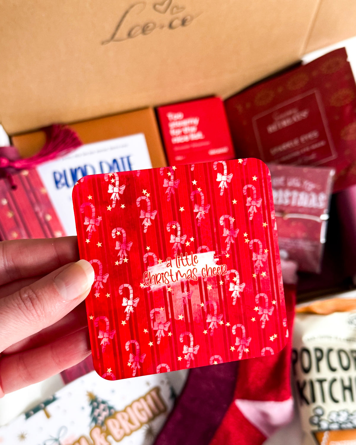 A hand holds a square red coaster with a festive pattern of candy canes, stars, and the phrase 'a little christmas cheer' in gold. The background shows an open gift box filled with various Christmas themed items.