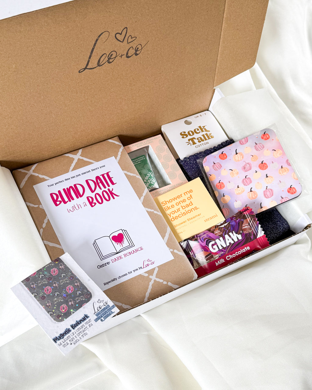 A curated gift box from Lee & Co containing a 'Blind Date with a Book' book, a sock talk tin, a patterned notebook, and a chocolate bar, arranged on a white fabric background.
