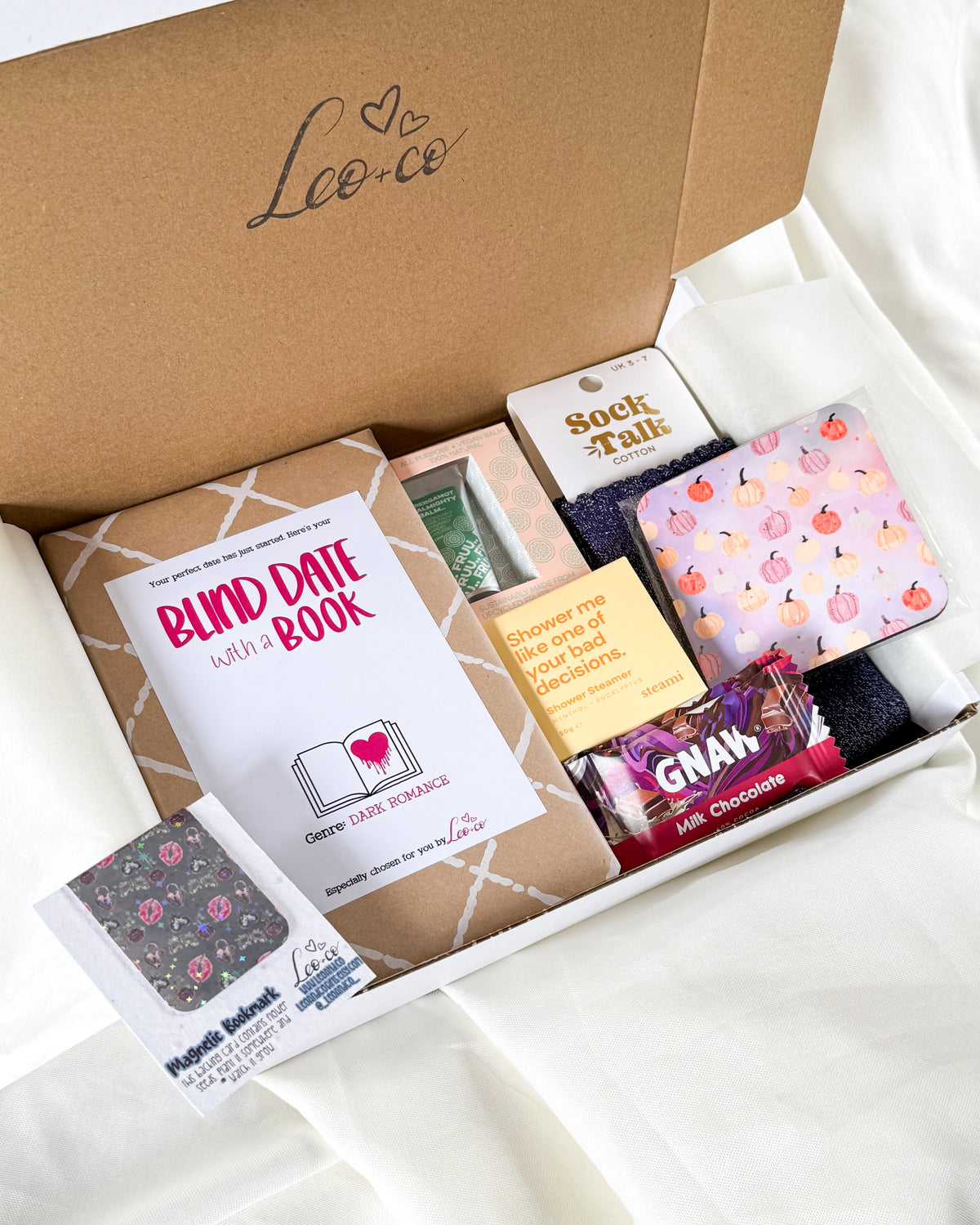 A curated gift box from Lee & Co containing a 'Blind Date with a Book' book, a sock talk tin, a patterned notebook, and a chocolate bar, arranged on a white fabric background.
