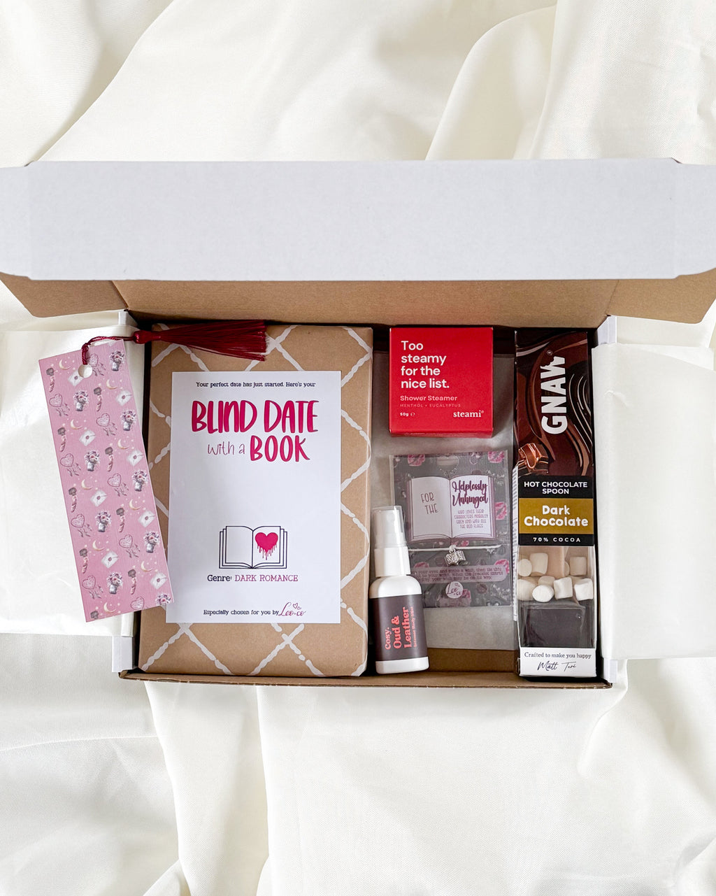 An open gift box arranged on white fabric, containing a pink patterned card, a book titled 'BLIND DATE with a BOOK', a small bottle, a boxed item labeled 'Two Scenery for the Day Book', and a dark brown rectangular product box with 'GNAN' visible.