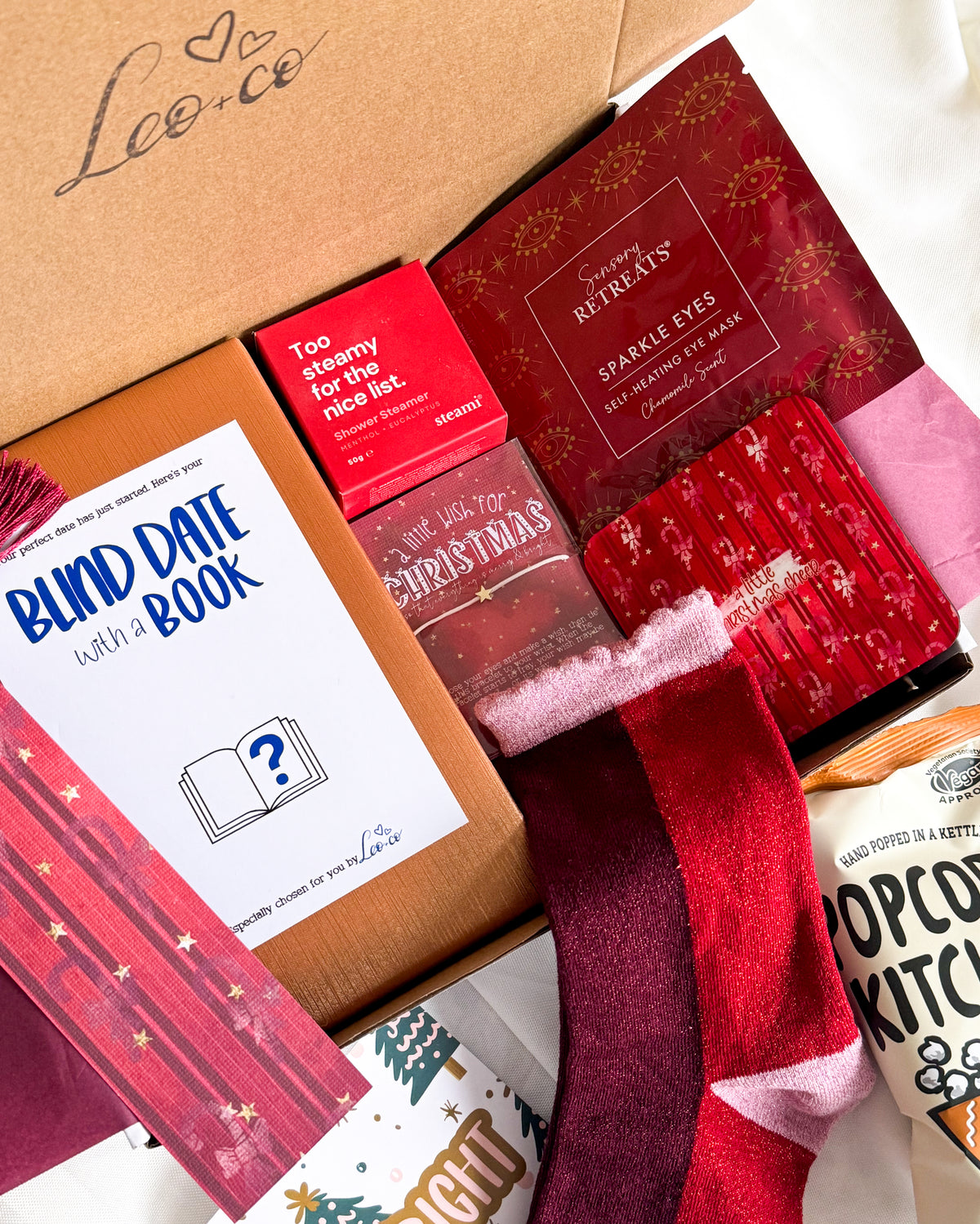 Christmas Blind Date With a Book gift box by Leo + Co, featuring a mystery book, cosy socks, shower steamer, Christmas wish bracelet, festive coaster, bookmark, self-heating eye mask and vegan gingerbread popcorn.
