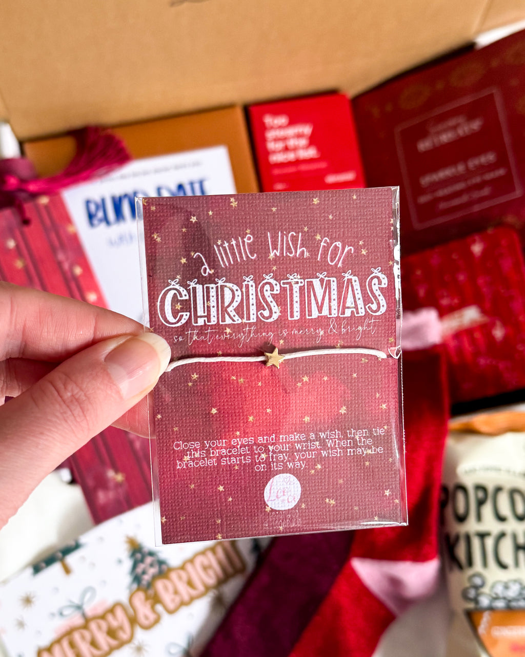 A hand holds a small red Christmas card that reads 'a little wish for CHRISTMAS' inside an open gift box with other packaged items around it.