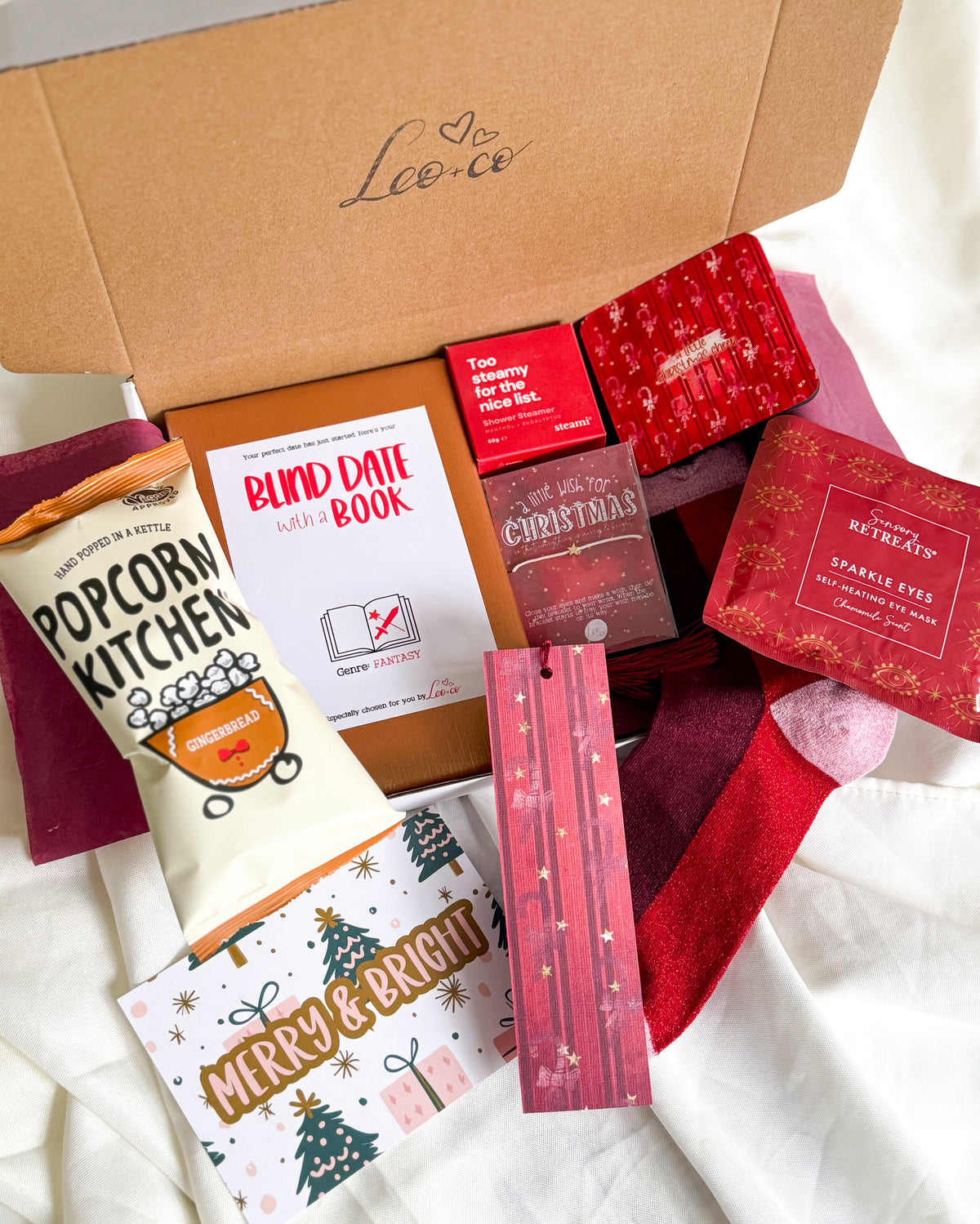 Christmas Blind Date With a Book gift box by Leo + Co, featuring a mystery book, cosy socks, shower steamer, Christmas wish bracelet, festive coaster, bookmark, self-heating eye mask and vegan gingerbread popcorn.