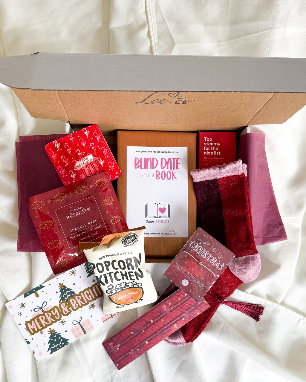 Christmas Blind Date With a Book gift box by Leo + Co, featuring a mystery book, cosy socks, shower steamer, Christmas wish bracelet, festive coaster, bookmark, self-heating eye mask and vegan gingerbread popcorn.