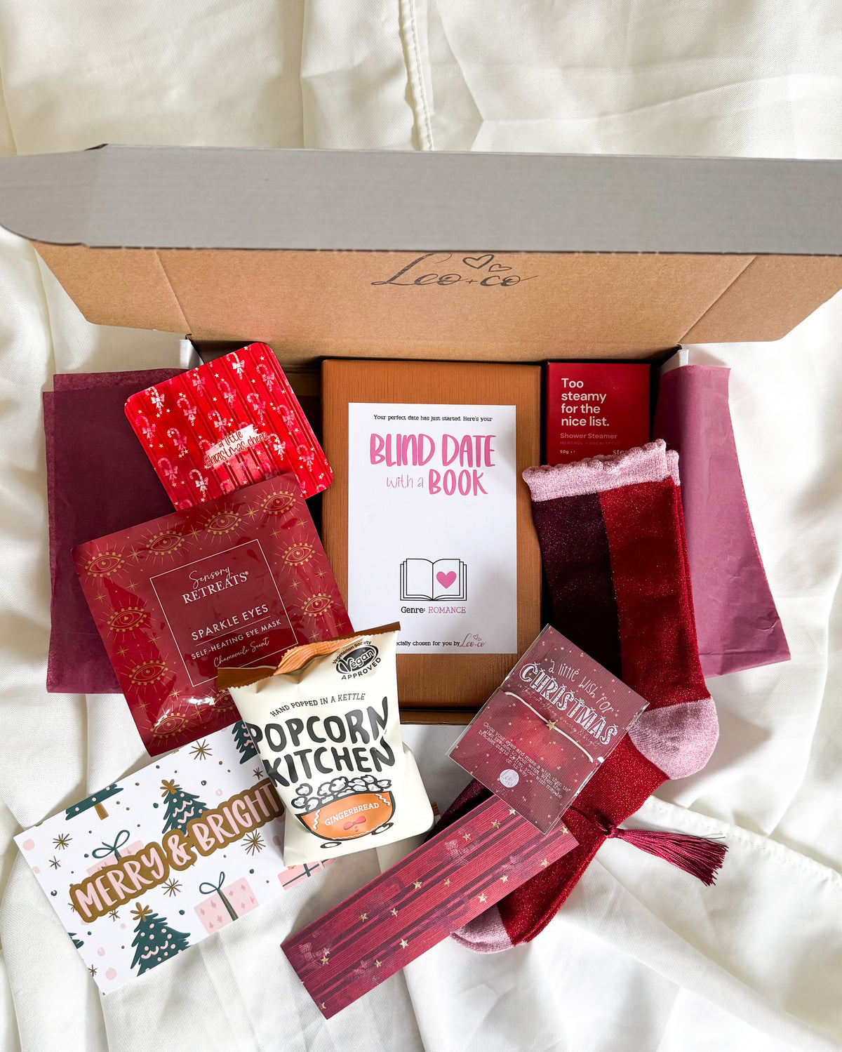 Christmas Blind Date With a Book gift box by Leo + Co, featuring a mystery book, cosy socks, shower steamer, Christmas wish bracelet, festive coaster, bookmark, self-heating eye mask and vegan gingerbread popcorn.