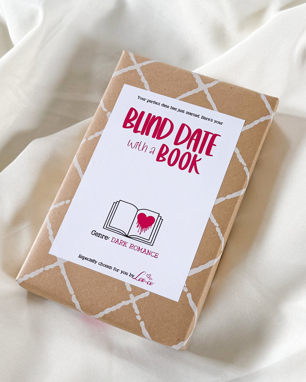 A wrapped book package on draped white fabric, featuring a kraft-paper cover with a white geometric line pattern and a large white label that reads 'BLIND DATE with a BOOK' in pink and black text. The label also includes the brand name 'Clear' and 'DARK ROMANCE'.