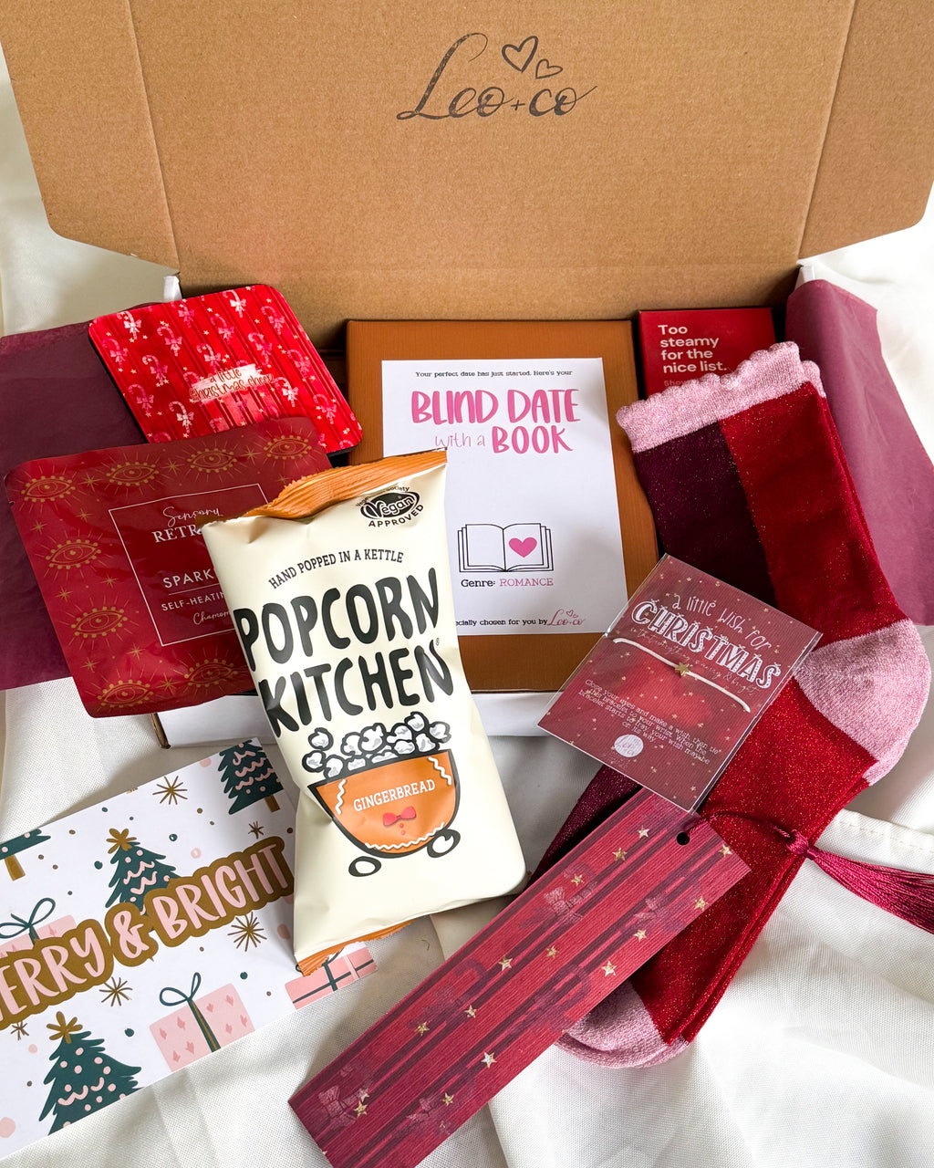 Christmas Blind Date With a Book gift box by Leo + Co, featuring a mystery book, cosy socks, shower steamer, Christmas wish bracelet, festive coaster, bookmark, self-heating eye mask and vegan gingerbread popcorn.
