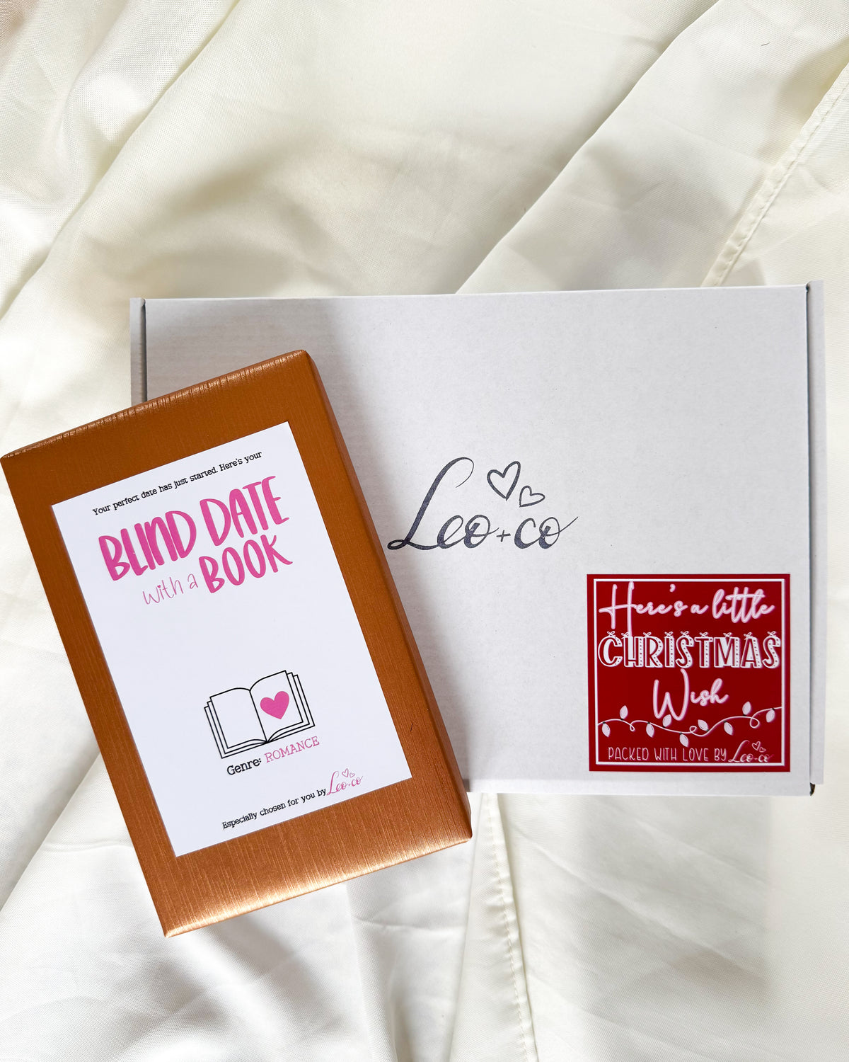 A gift set featuring a rose gold hardcover book labeled 'Blind Date with a Book' and a white gift box with 'Leo+Co' branding and a red 'Here's a little Christmas Week' sticker, arranged on a white fabric background.