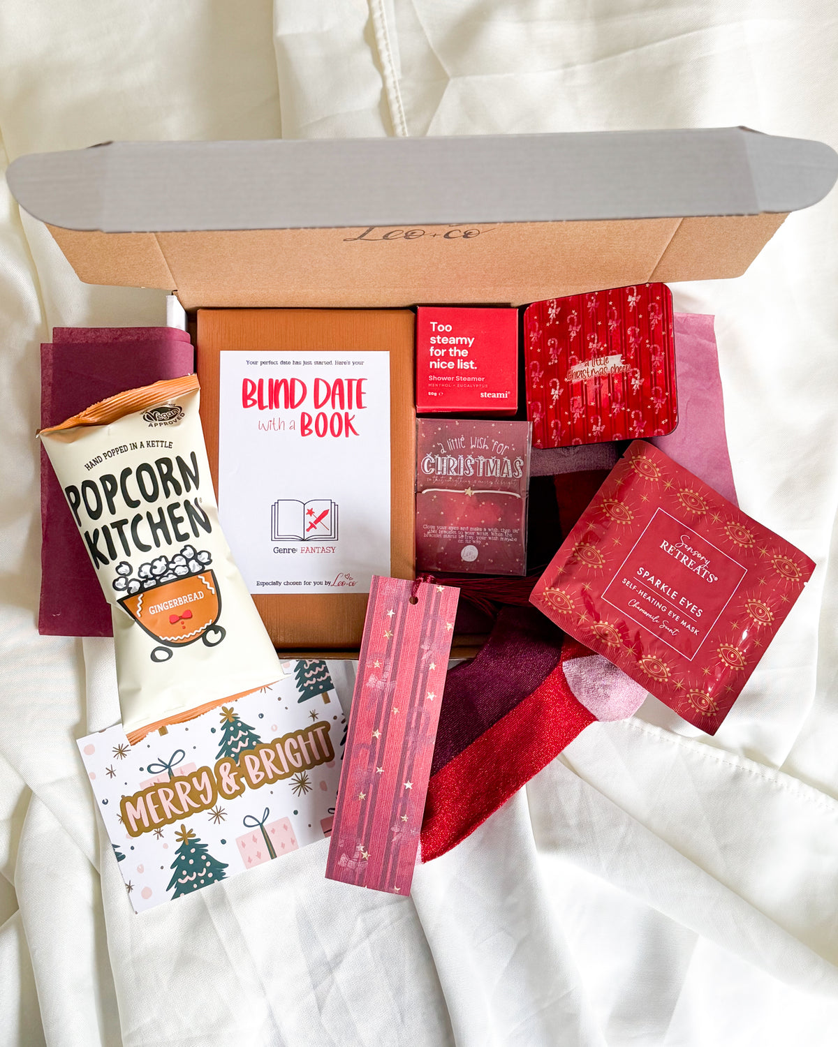 Christmas Blind Date With a Book gift box by Leo + Co, featuring a mystery book, cosy socks, shower steamer, Christmas wish bracelet, festive coaster, bookmark, self-heating eye mask and vegan gingerbread popcorn.