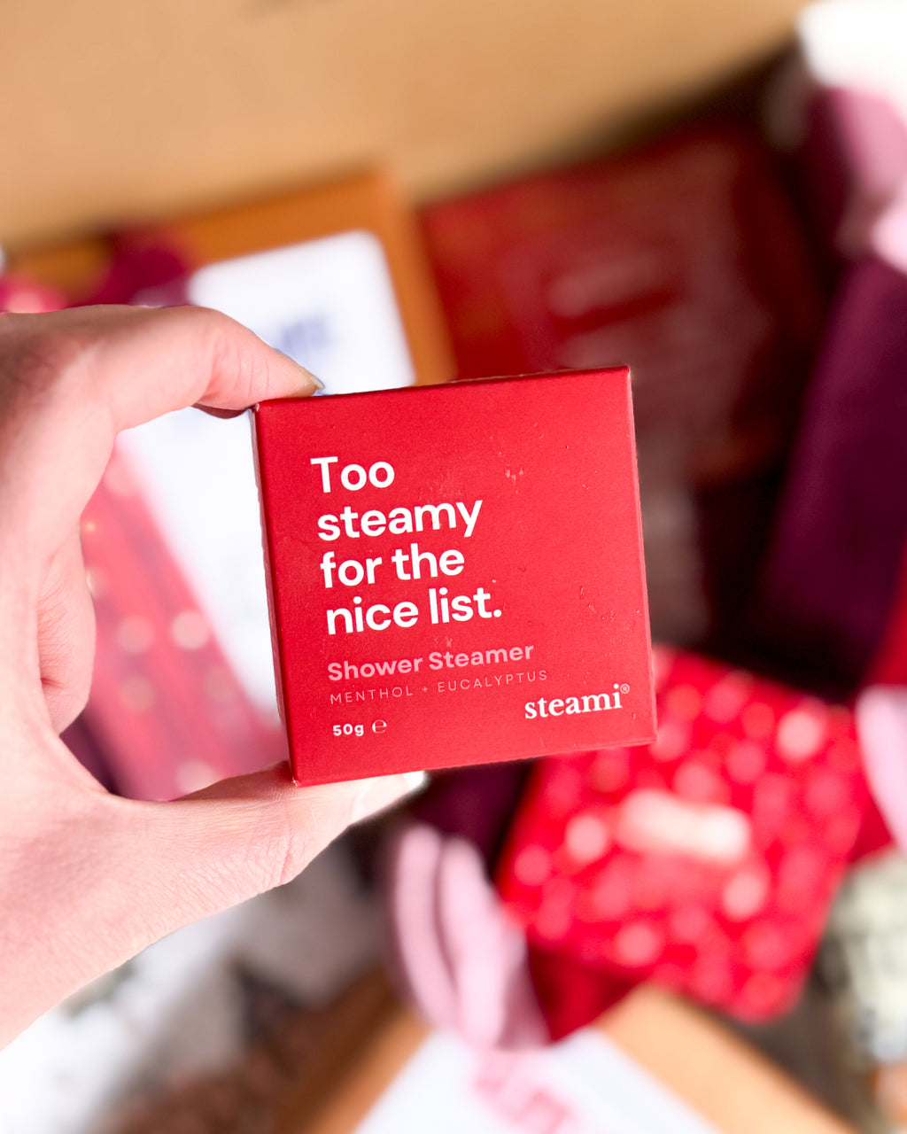 A hand holding a red square box of steami shower steamer, with the text 'Too steamy for the nice list. Shower Steamer MENTHOL + EXFOLIANTS 50g' visible, against a blurred background of red and pink items.