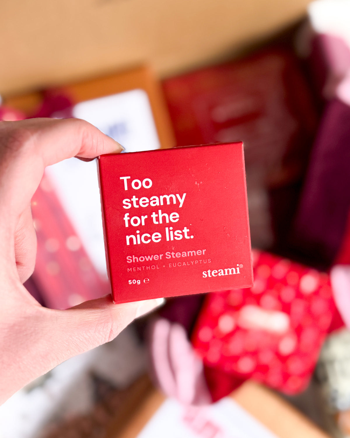 A hand holding a red square box of steami shower steamer, with the text 'Too steamy for the nice list. Shower Steamer MENTHOL + EXFOLIANTS 50g' visible, against a blurred background of red and pink items.