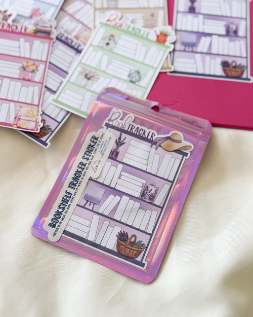 A packaged planner sticker set labeled 'Bookshelf Tracker Sticker' with a purple holographic sleeve, placed on top of several loose planner stickers featuring bookshelf illustrations and stationery-themed layouts on a cream fabric background.
