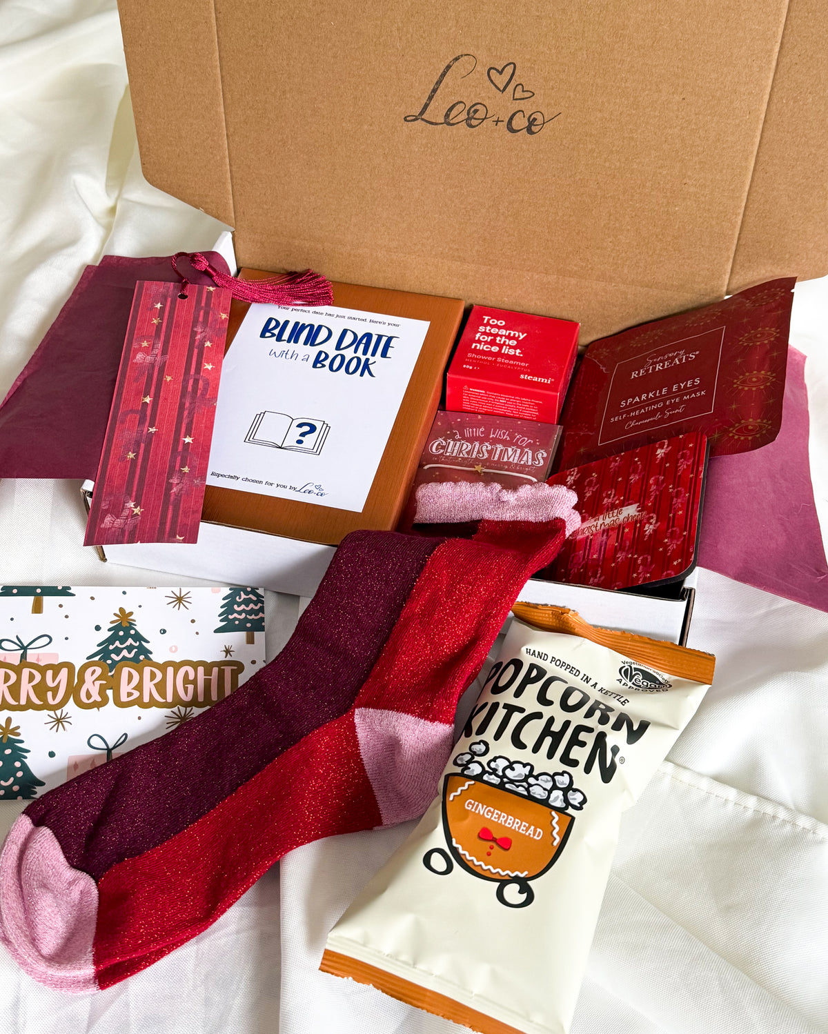 Christmas Blind Date With a Book gift box by Leo + Co, featuring a mystery book, cosy socks, shower steamer, Christmas wish bracelet, festive coaster, bookmark, self-heating eye mask and vegan gingerbread popcorn.