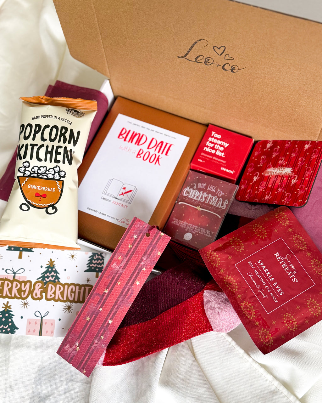Christmas Blind Date With a Book gift box by Leo + Co, featuring a mystery book, cosy socks, shower steamer, Christmas wish bracelet, festive coaster, bookmark, self-heating eye mask and vegan gingerbread popcorn.