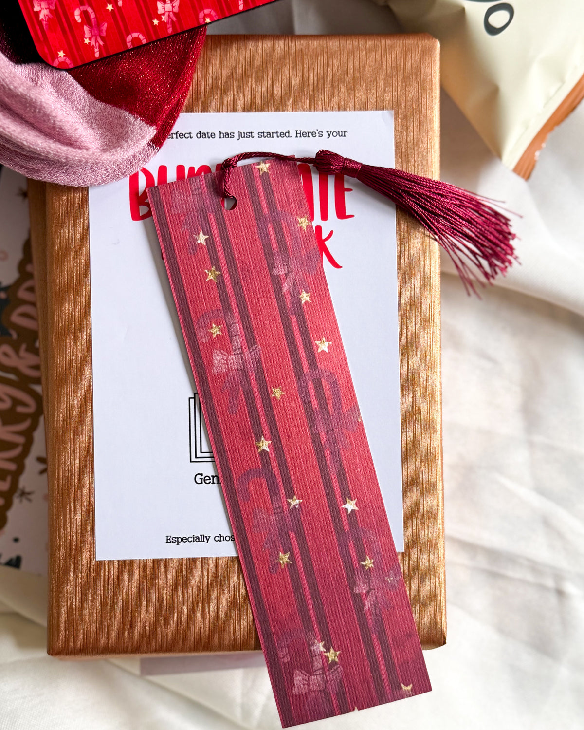 A red rectangular bookmark with gold stars and a matching tassel is laid across a Christmas Blind Date With A Book gift box and white card.