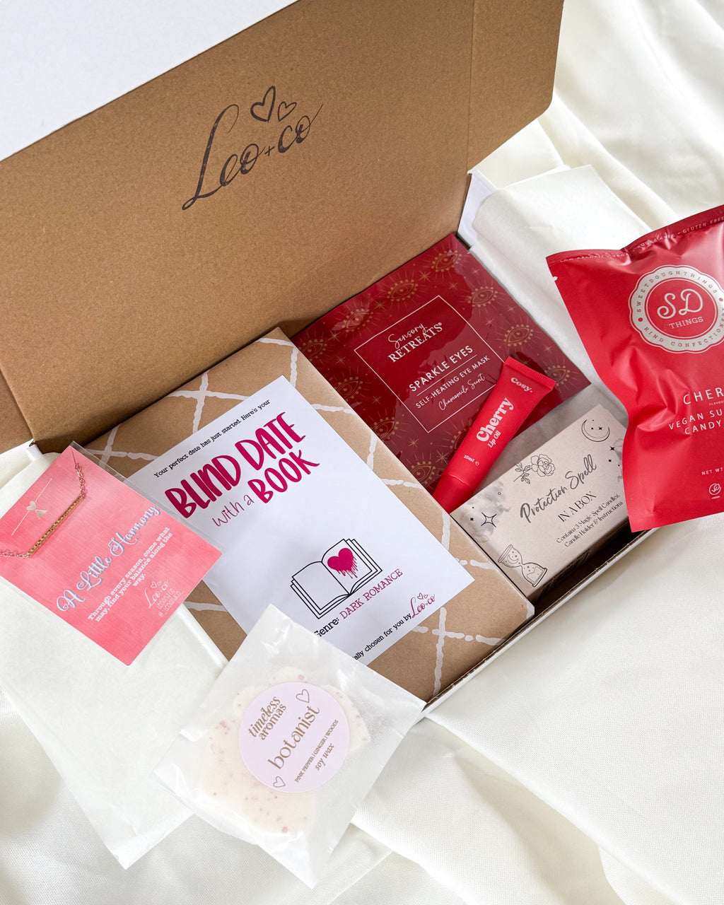 An open brown gift box filled with curated self-care and beauty items on white fabric. Visible contents include a carded book titled 'BNDY DATE BOOK', red packaged items, a pink packet, a small boxed item labeled 'Mister Soft', a clear bag with a round pink item, and a white pouch.