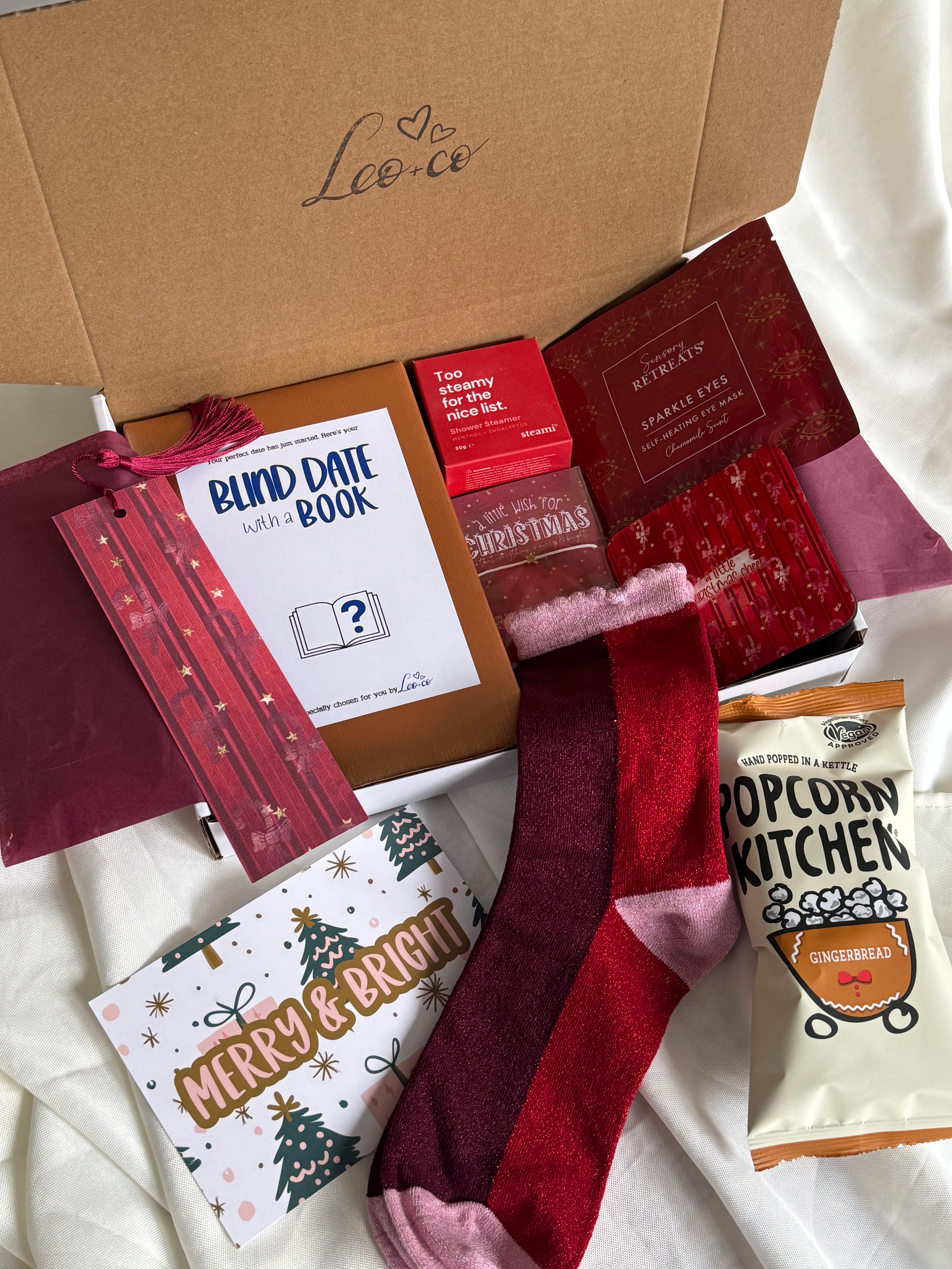 Christmas Blind Date With a Book gift box by Leo + Co, featuring a mystery book, cosy socks, shower steamer, Christmas wish bracelet, festive coaster, bookmark, self-heating eye mask and vegan gingerbread popcorn.